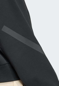 Black sleeve with a diagonal dark gray reflective stripe detail, paired with beige pants against a light gray background.