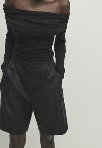 Haut noir à épaules dénudées à manches longues associé à un short noir ajusté avec un bouton à l'avant et une texture lisse.