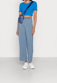 Modrý crop top, široké kalhoty s vzorem v světle modré barvě s geometrickými designy, doplněné malou modrou taškou a bílými teniskami.