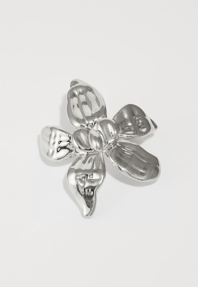 Sudraba ziedu piespraude ar noapaļotiem ziedlapām, mainīgām tekstūrām un pulētu apdari. Centrā ir ziedu pumpuru grupa, kas atspoguļo gaismu.