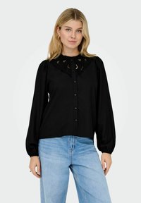 Donna bionda che indossa una camicetta nera con bottoni, dettagli in pizzo e maniche a sbuffo, abbinata a jeans azzurri contro uno sfondo bianco semplice.