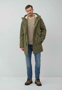 Groene, gevoerde jas met capuchon van zachte stof, met een fuzzy binnenkant, voorzakken en een knoopsluiting, gedragen over een beige trui en blauwe spijkerbroek.