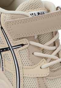 Zapatilla beige con tela de malla, cordones blancos, una correa de velcro y detalles de rayas laterales en blanco y negro.