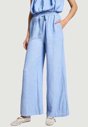 Femme portant un pantalon ample à jambes larges bleu clair texturé avec une taille à cordon et des chaussures plates blanches tissées sur un fond uni.
