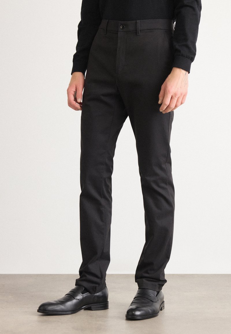 Zwarte broeken met een slim fit, voorzien van een riemlus tailleband, enkele knoopsluiting en een gladde textuur. Gecombineerd met zwarte leren schoenen.