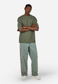 Olive žalios spalvos išplėstas marškinėliai su trumpomis rankovėmis, derinami su šviesiai mėlynomis džinsomis su šoninėmis juostelėmis raudonos ir baltos spalvų. Baltos sportbačiai.