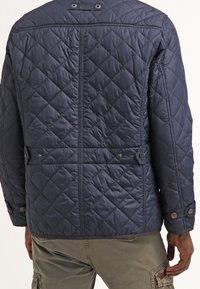 Mørkeblå quiltet jakke med diamantmønster, trykknapper og en opbygget ryg. Har justerbare taljejusteringer og sidelommer.