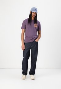 Purple short-sleeve t-shirt with orange graphic, dark blue loose-fitting denim jeans, light blue cap, and beige sneakers.