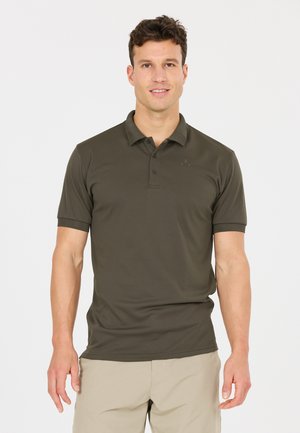 Mann, der ein dunkelgrünes kurzärmeliges Poloshirt und beige Hose trägt, steht vor einem einfarbigen hellen Hintergrund, blickt nach vorne und lächelt.