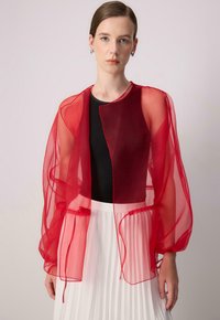 Rote durchsichtige Organza-Jacke mit leicht offenem Vorderteil und puffigen Ärmeln, über einem schwarzen Oberteil und einem weißen Faltenrock getragen.