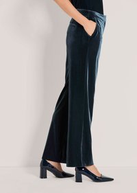 Vrouw in donkere fluwelen wijde broek en zwarte patenten block heel schoenen, staand met één hand in de zak.