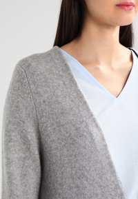 Ljusgrå ullcardigan med mjuk textur, med en sjalkrage och öppen framsida, lager på lager över en blekblå V-ringad tröja.