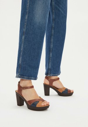 Stopy w niebieskich dżinsach i brązowo-beżowo-granatowych sandałach na platformie z odkrytymi palcami i paskami wokół kostek na białym tle.