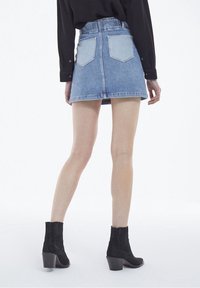 Persona que lleva una mini falda de mezclilla azul claro con bolsillos traseros, una camiseta negra de manga larga y botas negras de tacón hasta el tobillo, de pie sobre un fondo blanco.