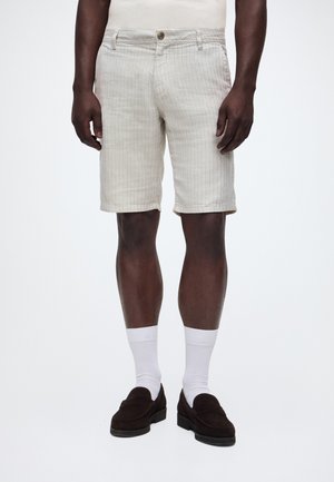 Homme portant un short beige rayé jusqu'aux genoux, des chaussettes blanches et des mocassins marron foncé, se tenant contre un fond clair uni.