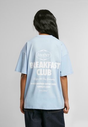 Lyseblå bomulds t-shirt med korte ærmer, med hvid tekst på ryggen: "Another Breakfast Club" og en henvisning til en café.