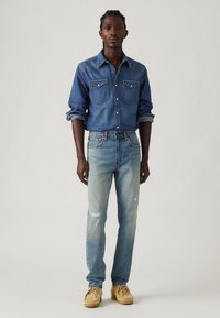 Cămașă din denim de culoare albastru închis, cu două buzunare pe piept și închidere cu nasturi, purtată alături de blugi drepți de culoare albastru deschis, uzati, și mocasini bej.