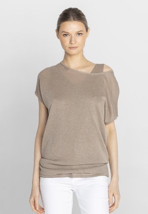 Młoda kobieta zwrócona twarzą do przodu, ubrana w błyszczącą taupe bluzkę odkrywającą ramiona i białe spodnie, stojąca na jednolitym jasnym tle.