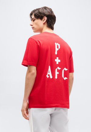 Νέος άνδρας με κόκκινο T-shirt που φέρει λευκά γοτθικά γράμματα "p + AFC" στην πλάτη, συνδυασμένο με ανοιχτόχρωμο παντελόνι, γυρισμένος προφίλ σε απλό φόντο.