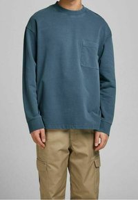 Person som bär en långärmad blå sweatshirt med rund hals och bröstficka tillsammans med beige cargobyxor mot en neutral bakgrund.