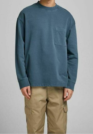 Henkilö, jolla on pitkähihaisen sinisen crewneck-sweaterin, jossa on rintatasku, ja vaaleat cargo-housut, taustalla on yksinkertainen tausta.