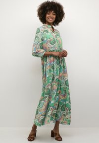 Dolga, zelena obleka s paisley vzorcem in tremi četrtinami rokavov, ovratnikom, poudarjenim pasom in sproščenim krilom. Oblečena z rjavimi sandali na blok peto.