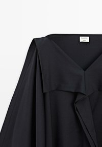 Czarna odzież z szerokim, płaskim kołnierzem i skośnym drapowaniem. Gładka tekstura, z minimalną ilością szwów i metką "Massimo Dutti Studio."