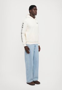 Cream pullover hoodie with front pocket, large logo on sleeve, paired with light blue baggy jeans and brown shoes. Simple design, relaxed fit.