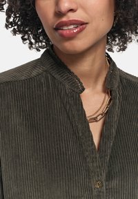Camicia in corduroy verde scuro con colletto alto, caratterizzata da una fine trama a coste e un dettaglio con bottoni. Accessoriata con collane d'oro sovrapposte.