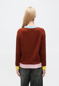Maglione lavorato a maglia color ruggine con scollo azzurro chiaro, bordi rosa e gialli. Tessuto strutturato e vestibilità comoda. Indossato con pantaloni grigi a vita alta.