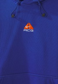 Felpa blu con una texture liscia, caratterizzata da un logo ACG arancione e da un'emblema Nike. Include cordini regolabili sul cappuccio.