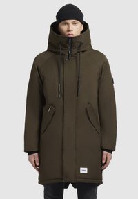 Parka verde măsliniu cu guler înalt, șnururi ajustabile, fermoar frontal și buzunare laterale. Dispune de o etichetă logo pe partea de jos din față.
