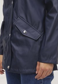 Närbild av person som bär en marinblå vattentät jacka med tryckknappsfickor och blå jeans, handen vilande vid sidan.