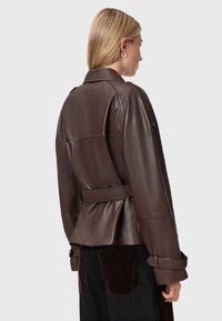 Bruine leren jas met een ceintuur in de taille en gestructureerde mouwen. Heeft een gladde textuur en een licht uitzwenkende achterzoom.