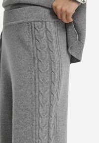 Pull en tricot gris avec un motif en relief sur le côté, des poignets côtelés et une texture douce. Gros plan sur le détail du tissu.