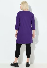 Lila tunika med trekvartslånga ärmar, gjord av mjukt tyg, i kombination med svarta leggings och svarta plattformsskor. Slät textur, avslappnad design.