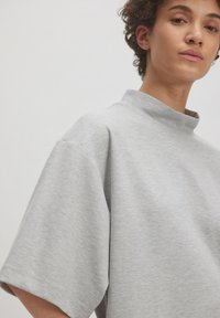 Lichtgrijze oversized t-shirt met een hoge hals en korte mouwen, gemaakt van zachte, gestructureerde stof. Heeft een relaxte pasvorm en een minimalistisch ontwerp.