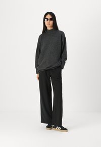 Mørk grå oversized genser med høy nakke, kombinert med vide svarte bukser og svarte sneakers med hvite striper. Tilbehøret inkluderer solbriller.