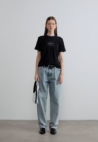 Μαύρο βαμβακερό μπλουζάκι με λογότυπο, ανοιχτό μπλε τζιν παντελόνι, μαύρα παπούτσια, κρατώντας ασημί τσάντα. Απλό, καθημερινό ντύσιμο σε μινιμαλιστικό περιβάλλον.