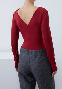 Punainen pitkähiha bodysuit, jossa on matala V-pääntie selässä, yhdistettynä harmaisiin teksturoituihin korkeavyötäröisiin housuihin. Sujuva kangasviimeistely.
