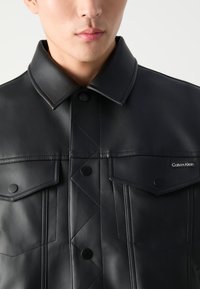 Čierna kožená bunda s prešívaným dizajnom, dvomi náprsními vreckami, cvokovými gombíkmi a golierom. Logo Calvin Klein na vrecku.
