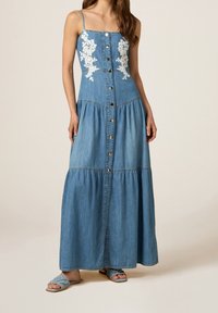 Robe maxi en denim avec des bretelles réglables, broderies florales, fermetures à boutons à l'avant et jupe à volants. Portée avec des sandales tressées bleues.