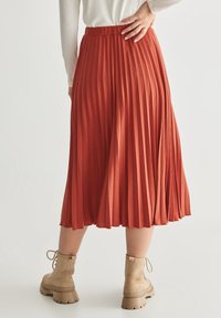 Faltenrock in rostfarbenem Midi-Format aus weichem Stoff. Verfügt über einen hohen Bund und reicht bis zur Mitte der Wade. Kombiniert mit hellbraunen Stiefeletten.