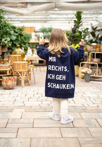 Navyblaues, übergroßes Sweatshirt mit weißem Text, getragen von einem Kind, kombiniert mit hellen Leggings und pastellfarbenen Sneakers, in einem Pflanzen gefüllten Raum.