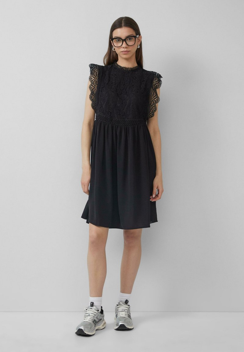 Robe noire avec un corsage en dentelle, des manches à volants et une taille froncée. Porte des chaussures de sport argentées et des chaussettes blanches, debout sur un fond uni.