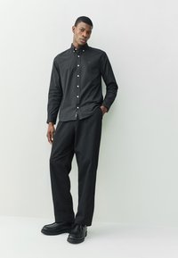 Camisa negra con botones y cuello, combinada con pantalones negros. Presenta un corte ajustado, tela texturizada y un pequeño detalle de logo bordado.