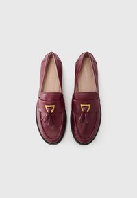 Burgundy leather loafers with a front tassel and gold accent. Smooth texture, rounded shape, featuring contrast stitching and a black sole.