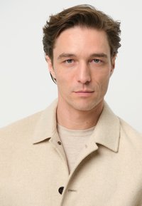 Giacca beige in misto lana con colletto a cappotto, chiusura a bottoni e tessuto testurizzato; indossata sopra una camicia grigia chiara.