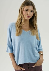 Svetlo modrý pletený sveter s výstrihom do V a rukávmi po trištvrte, s voľným strihom a jemnou textúrou. Skombinovaný s tmavými nohavicami.