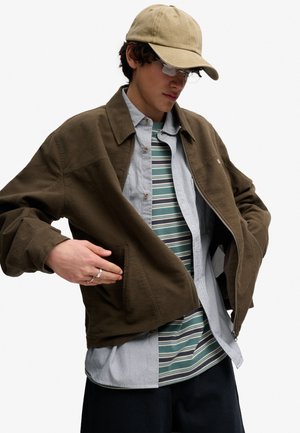 Młody mężczyzna w beżowej czapce, okularach, brązowej kurtce na paskowanej koszuli i overshirtcie, z rękami poprawiającymi kieszeń kurtki.